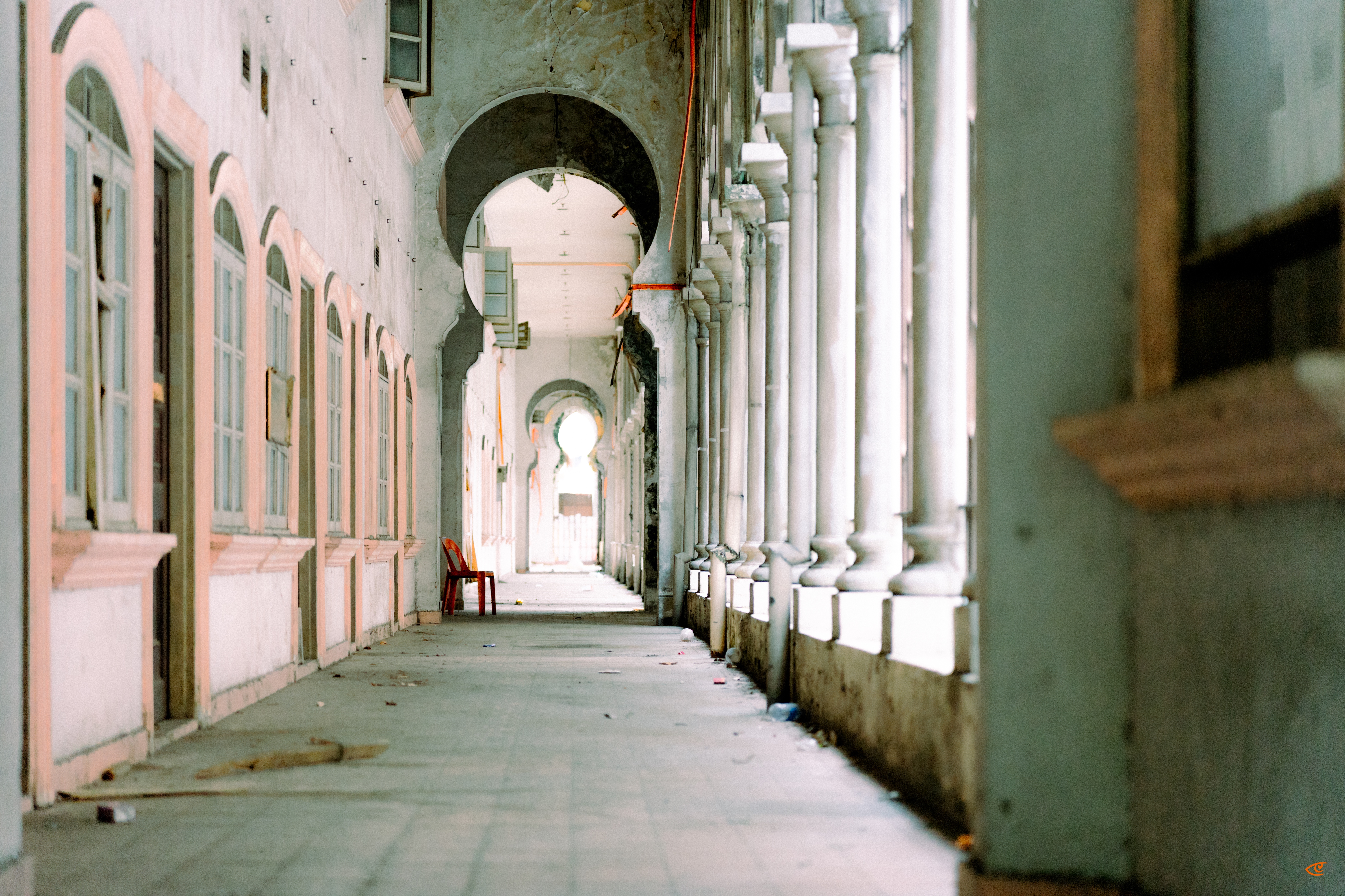 Long arched corridor with tiled floor, pastel pink walls and shuttered windows on the left, and a row of white columns on the right, leading to a bright archway in the distance; a red chair sits along the left wall and scattered debris lies on the floor.