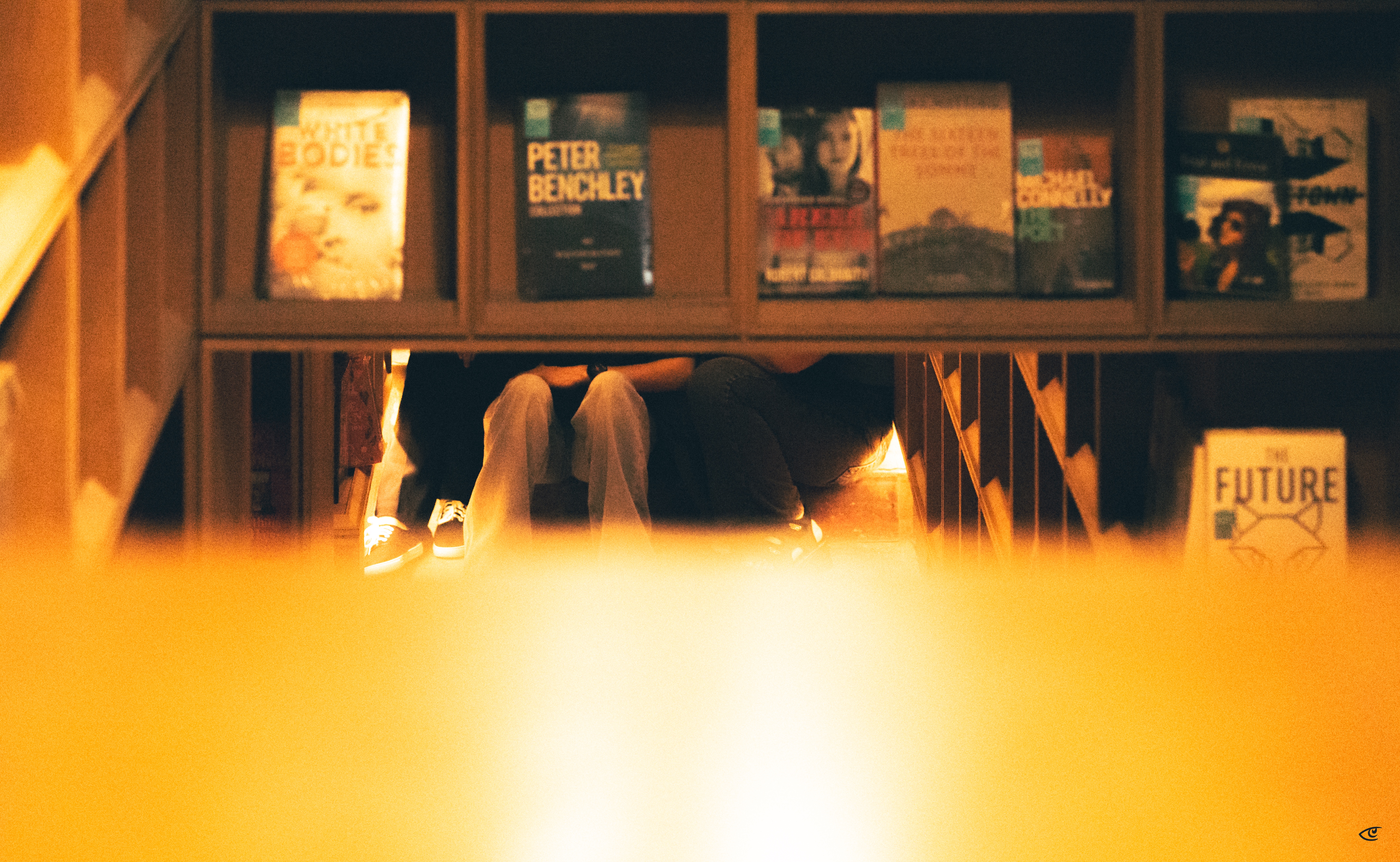 Low-angle view through a wooden bookshelf with book covers on the top shelf; a seated person’s bent knees and legs are visible in a narrow gap behind the shelf, with warm indoor lighting and blurred foreground.