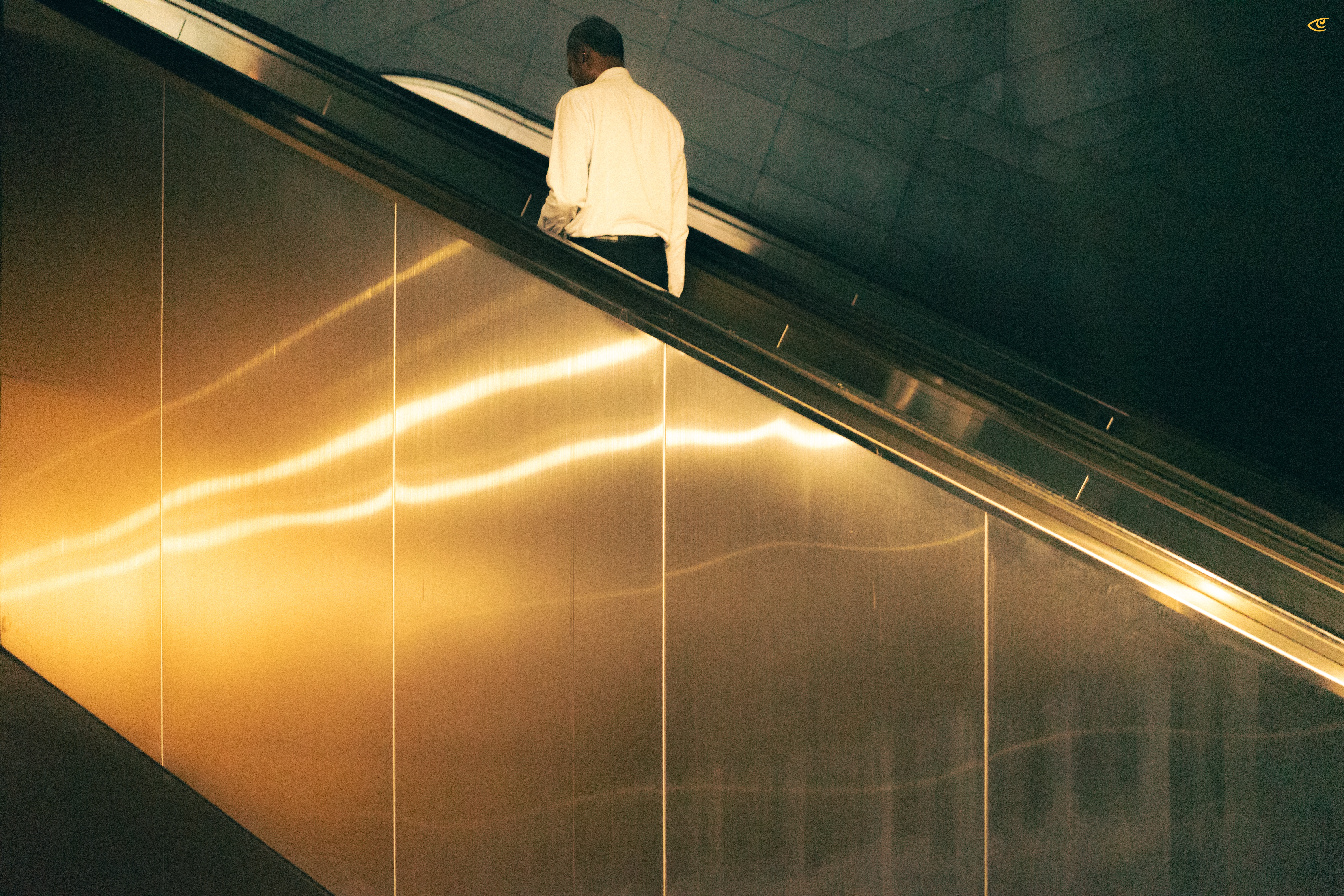 Man in a white shirt riding an escalator upward, viewed from the side, with reflective gold-toned wall panels and a tiled ceiling in the background.