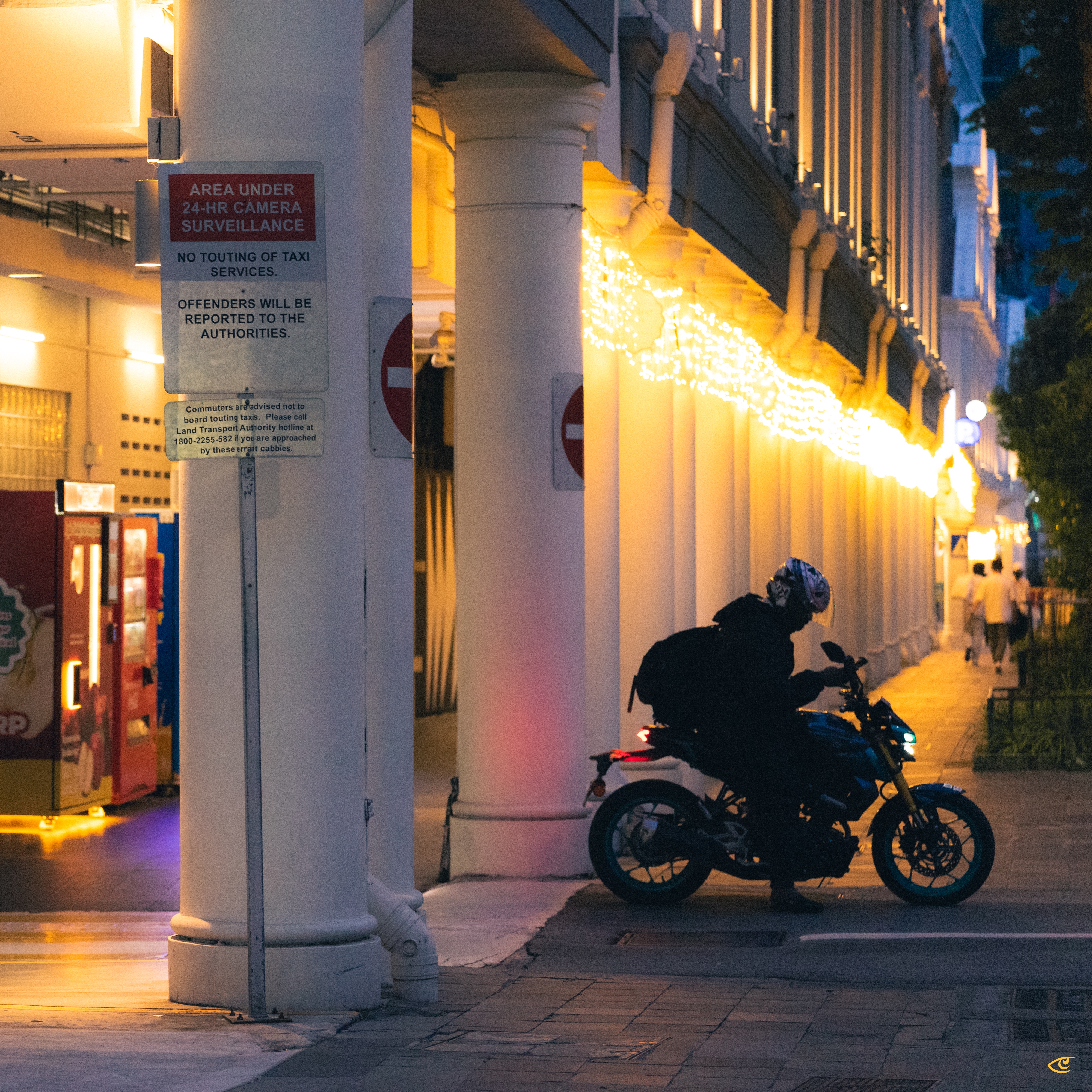 Motorcyclist wearing a helmet and backpack stopped at the curb beside a row of white columns under warm string lights at dusk; a “24-hr camera surveillance” sign and vending machines are on the left, with a lit sidewalk and a few pedestrians in the distance.