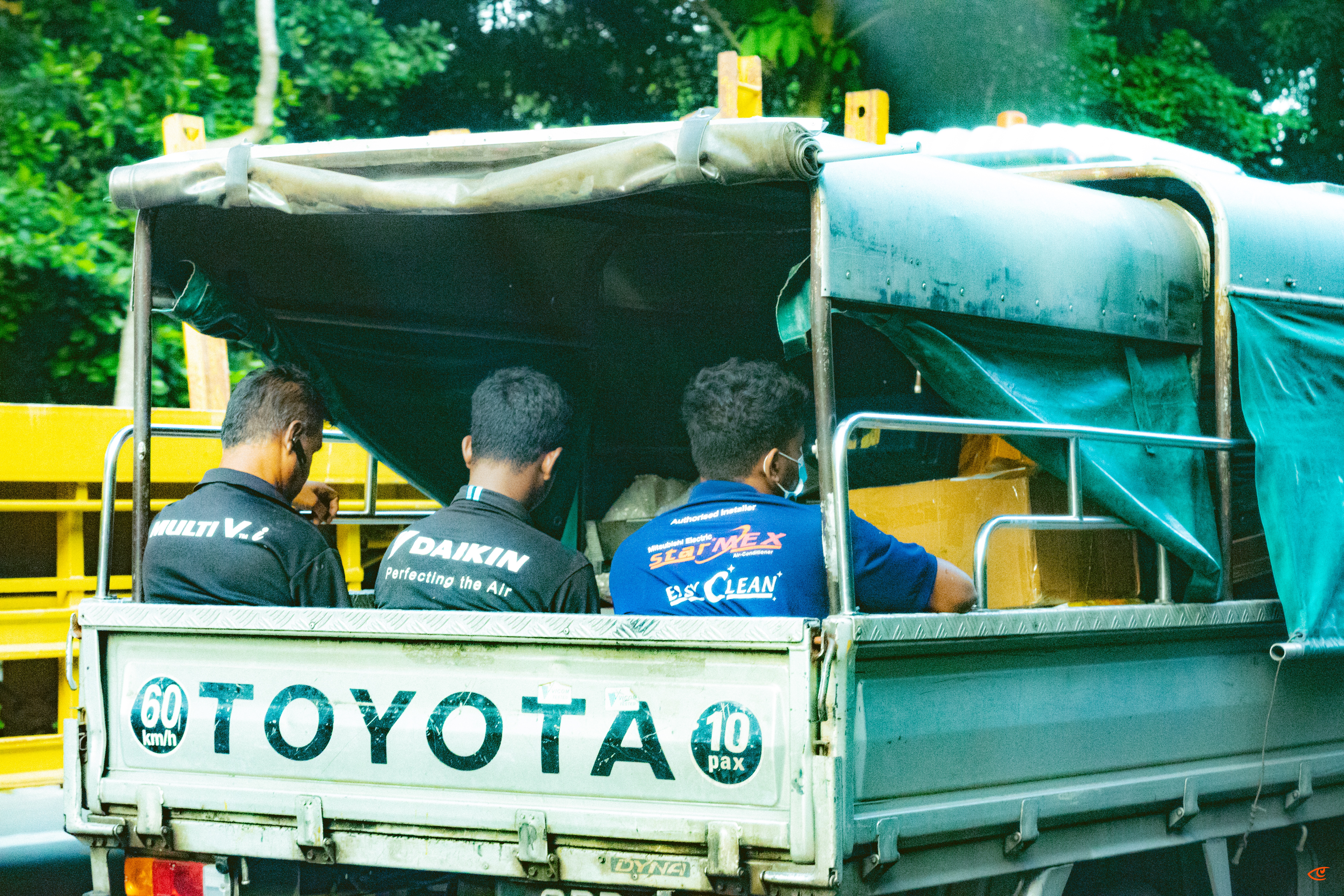 Three men seated with their backs to the camera in the open rear of a Toyota pickup truck, partially covered by a tarp canopy; “TOYOTA” printed on the tailgate with “60 km/h” and “10 pax” stickers, yellow railings and green trees in the background.