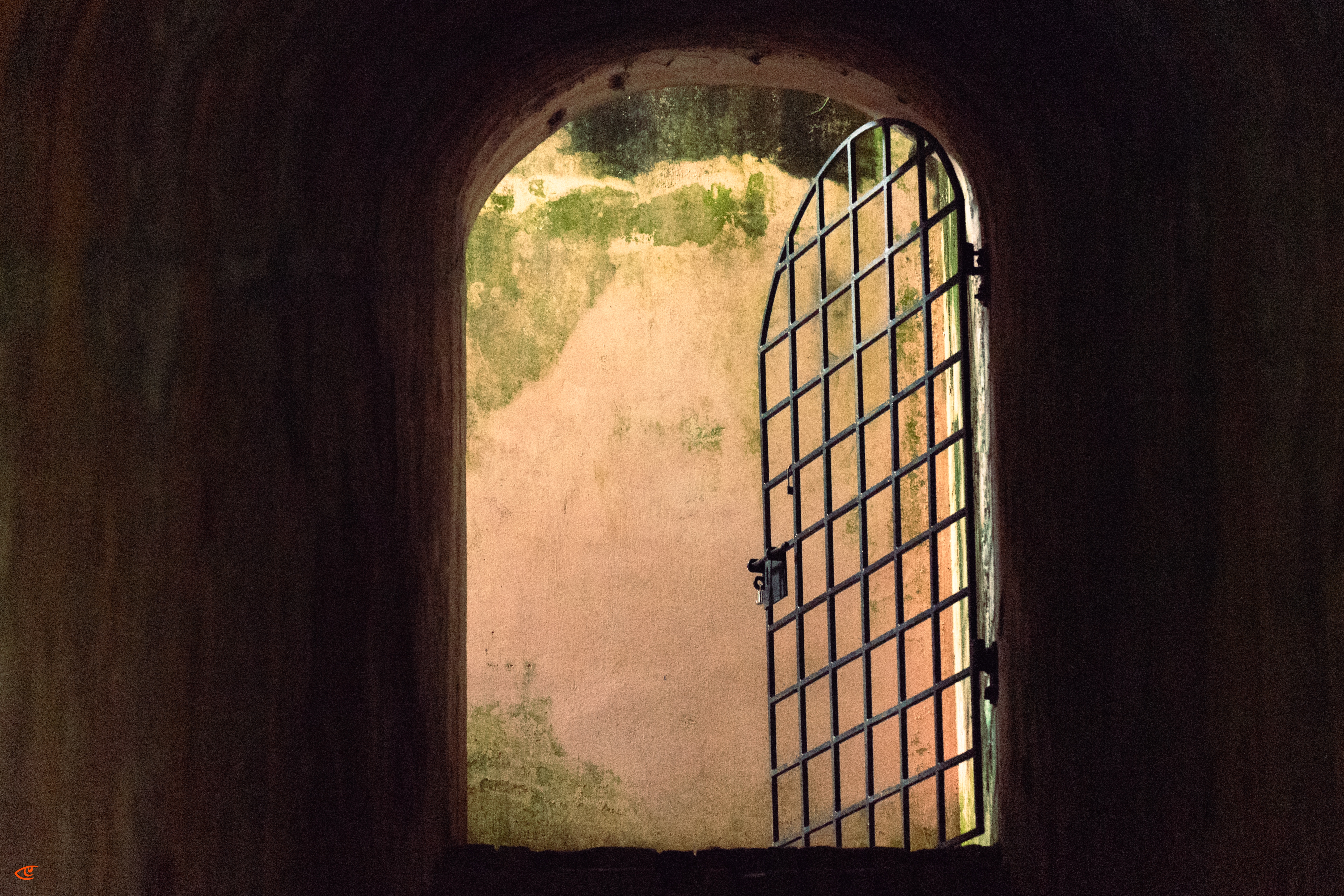 View from inside a dark arched tunnel toward a sunlit doorway, with a partially open iron-bar gate on the right and a weathered wall with peeling plaster and green stains beyond.