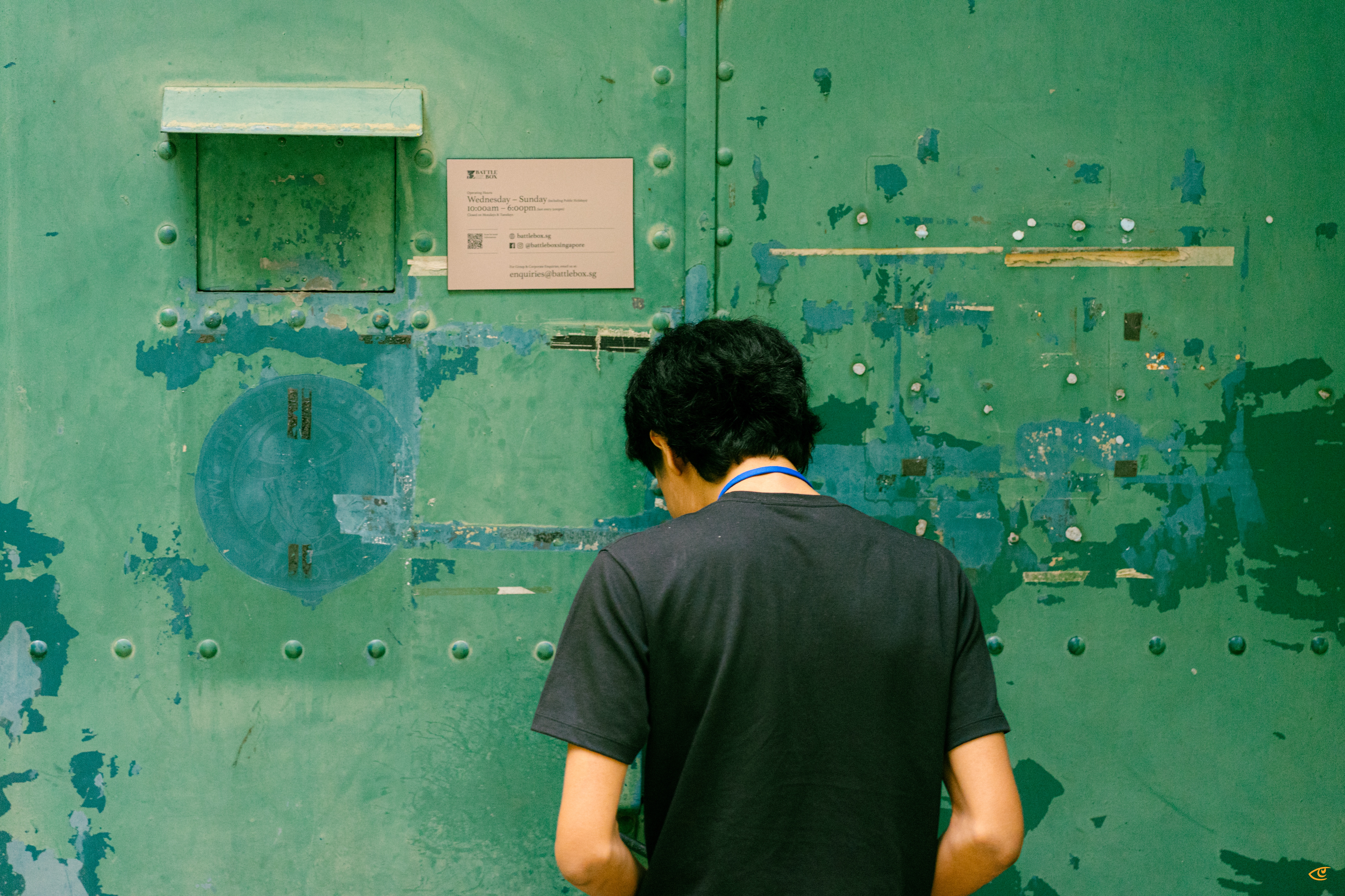 Person seen from behind wearing a dark T-shirt, standing against a weathered green metal door with peeling paint, rivets, a small shelf-like panel, and a posted notice.