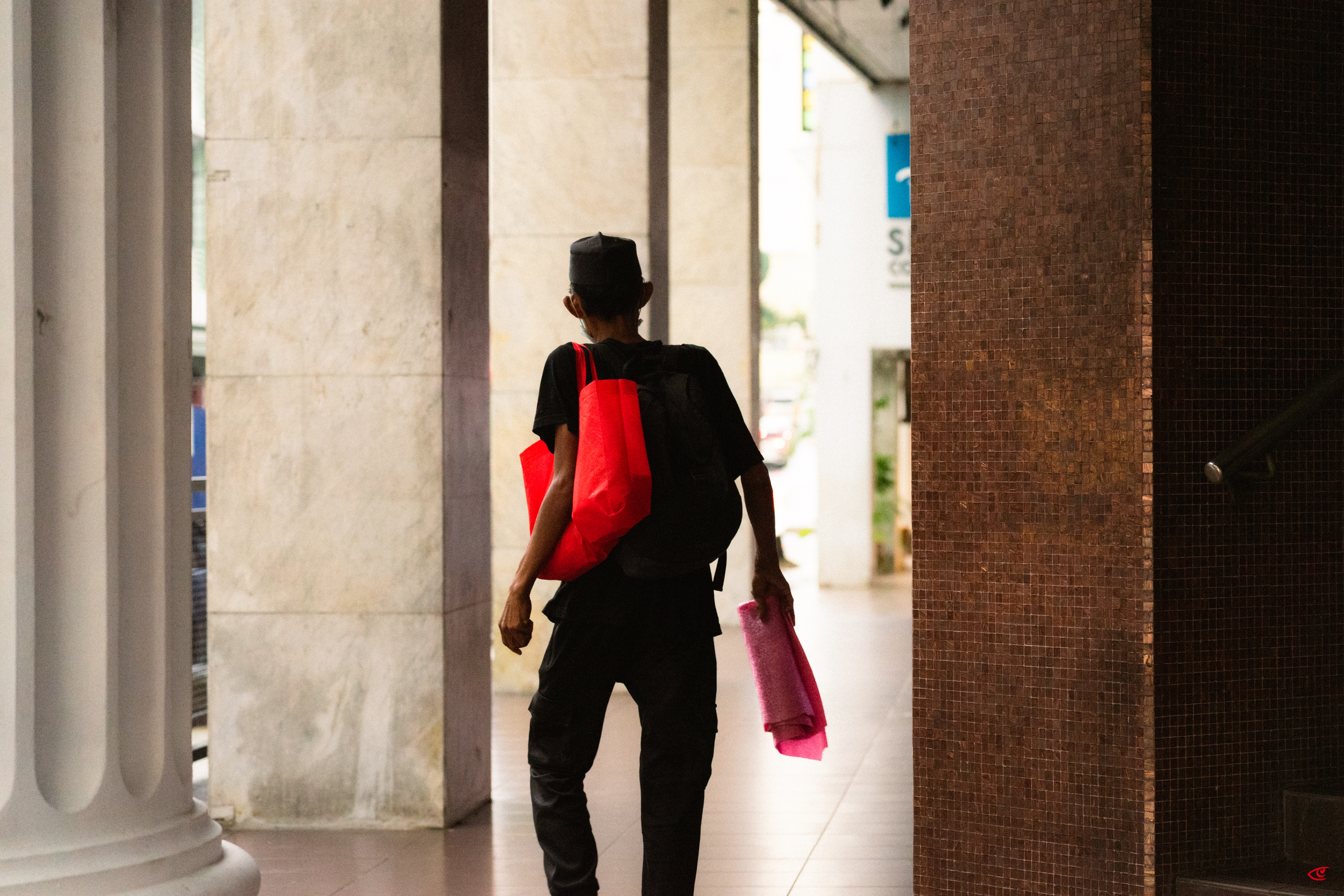 Person walking away through a covered walkway with tall columns, wearing black clothing and a cap, carrying a red tote bag and a black backpack, holding a pink item in one hand.