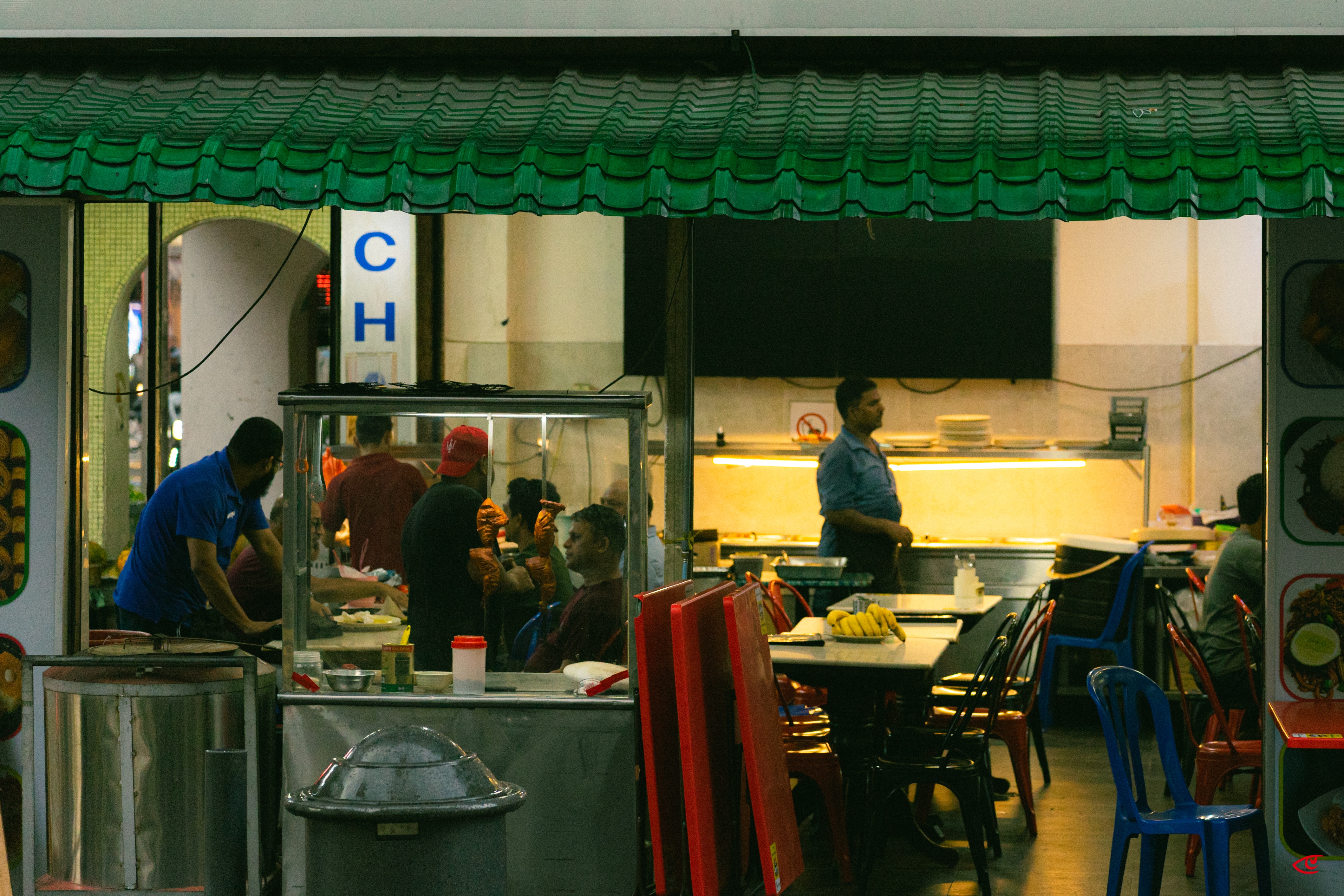 Street-side eatery with a green awning; workers prepare food at a glass-fronted stall on the left while diners sit at tables and plastic chairs on the right, with a lit counter and blank wall-mounted TV in the background.
